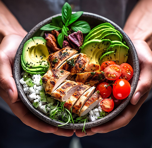 a person holding a dark ceramic bowl of grilled chicken, avocado, tomatoes and tofu